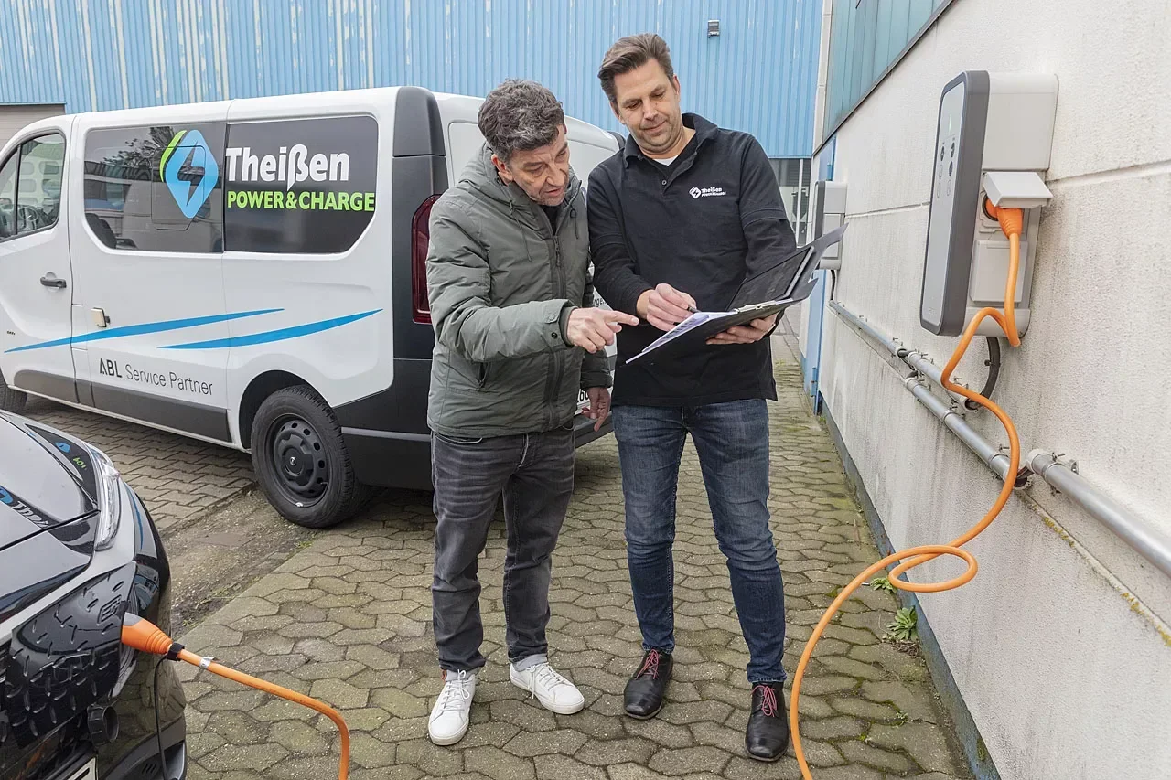 Zwei Techniker prüfen mit einem Laptop eine Wallbox-Ladestation, daneben ein Servicefahrzeug von Theißen Power & Charge